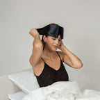 Woman wearing a black silk sleep mask in bed with white bedding.
