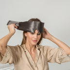 Woman wearing a dark gray silk eye mask with lace trim, holding it up to her face.