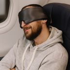 Man sleeping on an airplane with a gray hoodie and black sleep mask.