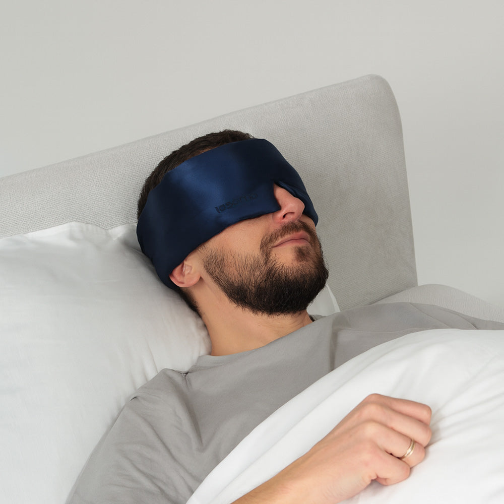 Man wearing a navy blue silk sleep mask while lying in bed with white bedding and pillows.