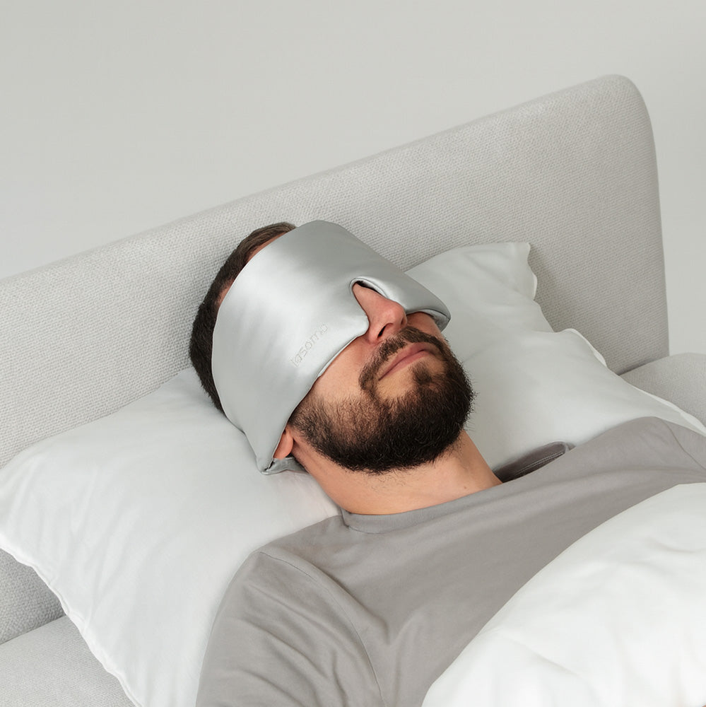 Man lying in bed wearing a silver silk sleep mask.