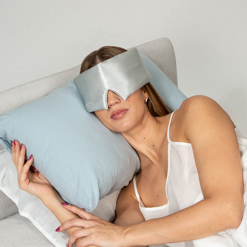 Woman lying on a bed with a blue pillow and wearing a silver silk sleep mask.